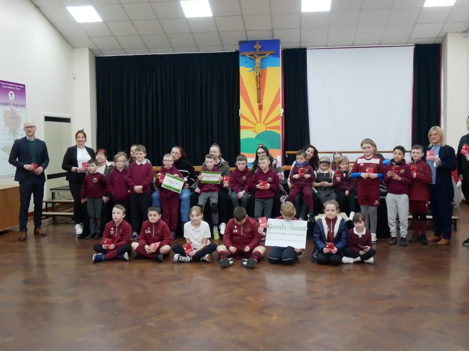 School pupils and their parents posing with goods that have been given to them by Goods for Good, The parents are sitting on a stage and the children sit on the floor in front of them. Stephen Kinnock MP stands on the left hand side of the photo. 